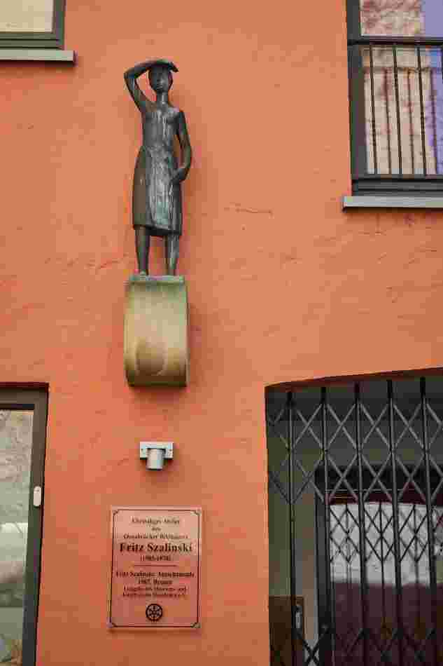 Auf einem hohen Sockel am Haus steht die kleine Bronzefigur „Ausschauende“. Sie hat ihre rechte Hand an die Stirn gelegt und schaut in die Ferne.