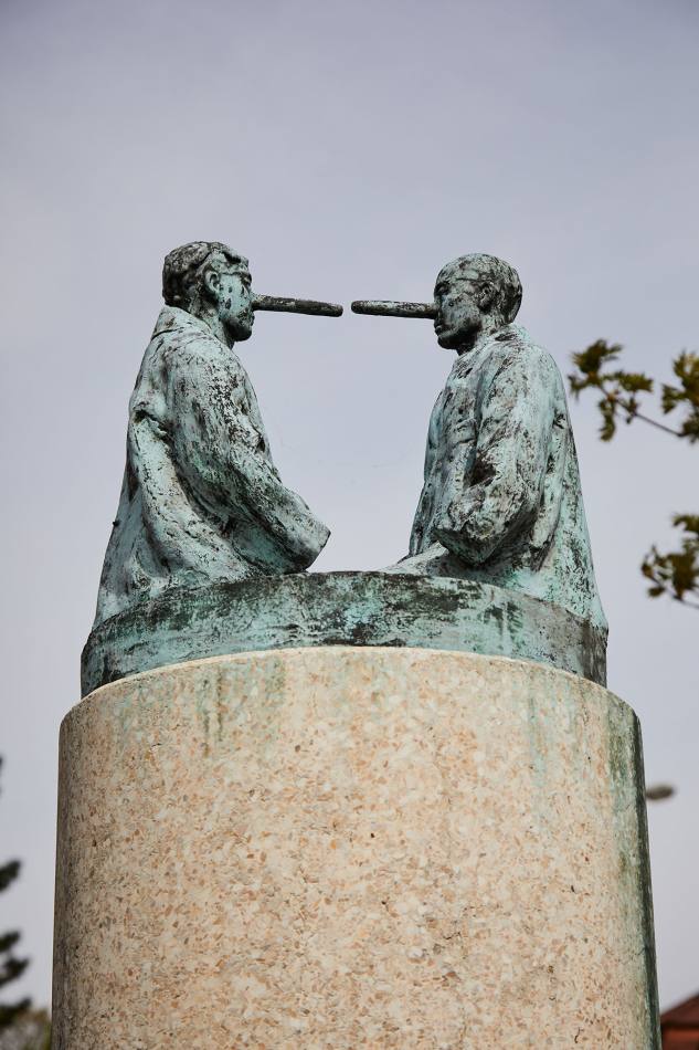 On a pillar are 2 men made of bronze. The two men are looking at each other. They have long noses, just like the figure of Pinocchio. They are wearing huge coats in which their hands disappear. They are kneeling. But you can't see exactly where their legs end. Because you can't see that and because their faces are so exaggerated, the sculpture looks strange and a little scary.