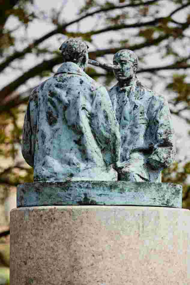 Auf einer Säule sind 2 Männer aus Bronze. Die beiden Männer schauen sich an. Sie haben lange Nasen, so wie bei der Figur Pinocchio. Sie tragen riesige Mäntel, in denen ihre Hände verschwinden. Sie knien. Aber man kann nicht genau sehen, wo ihre Beine enden. Weil man das nicht sehen kann und weil ihre Gesichter so übertrieben sind, sieht die Skulptur seltsam und ein bisschen unheimlich aus.