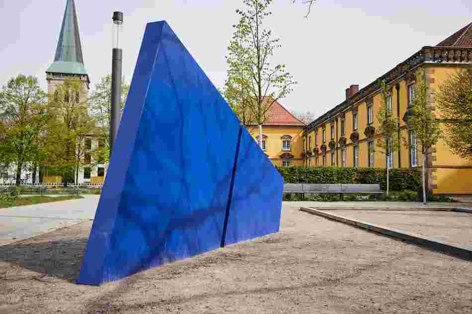 Vor dem Osnabrücker Schloss steht ein blaues Dreieck, das wie ein Zacken aus der Erde ragt und in der Mitte geteilt ist.