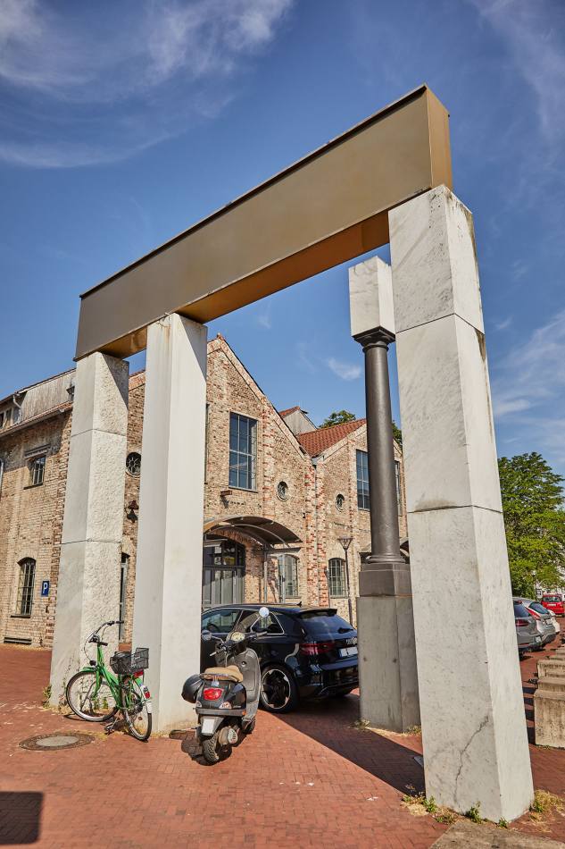 A portal consisting of three marble columns supporting a gold-painted steel beam. A fourth, black column stands next to it. A bicycle and a scooter are parked in front of the artwork. A car is parked behind the portal.