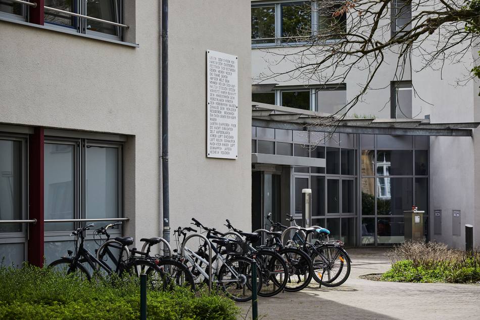 A large acrylic panel with words on it hangs on the wall next to the entrance to the town hall. There are many bicycles in front of the wall.