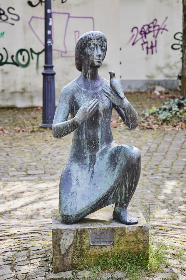Eine Bronzeskulptur auf einem niedrigen Sockel: Ein halb hockendes und halb kniendes Mädchen. Es hält einen Vogel in der linken Hand und die rechte Hand vor der Brust.