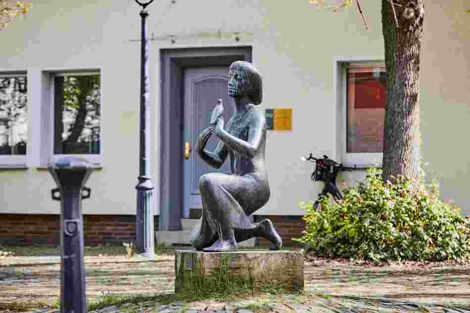 In der Mitte des Platzes steht eine kleine Bronzeskulptur auf einem niedrigen Sockel: Ein halb hockendes und halb kniendes Mädchen. Es hält einen Vogel in der linken Hand und die rechte Hand vor der Brust.