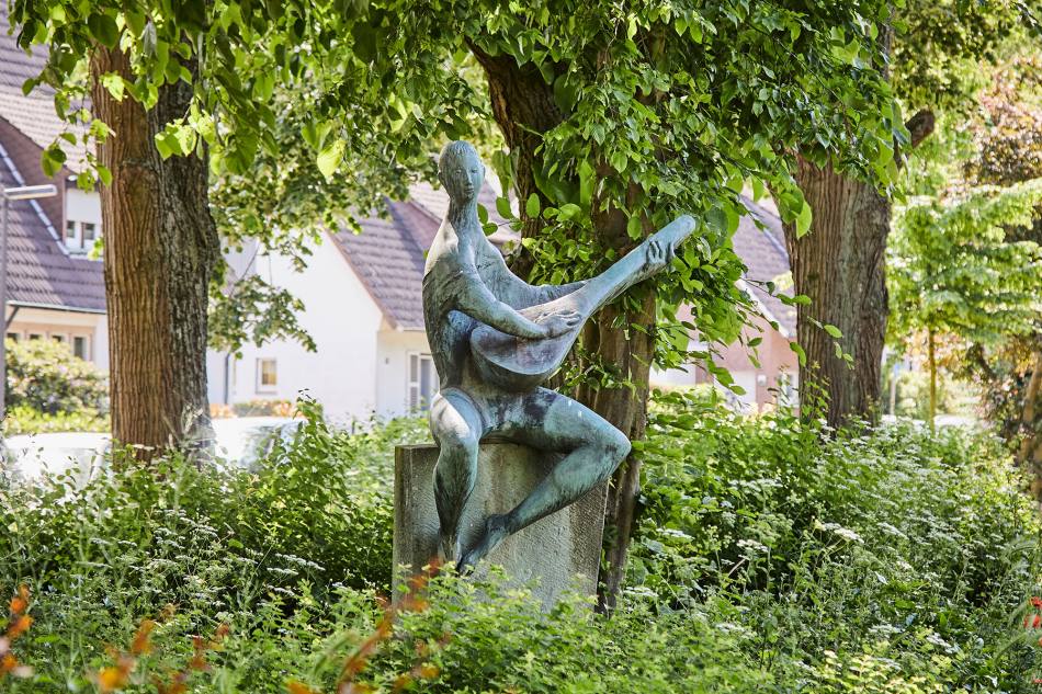 Eine männliche Figur aus Bronze sitzt auf einem Sockel und spielt die Laute. Hinter der Skulptur stehen 3 Bäume.