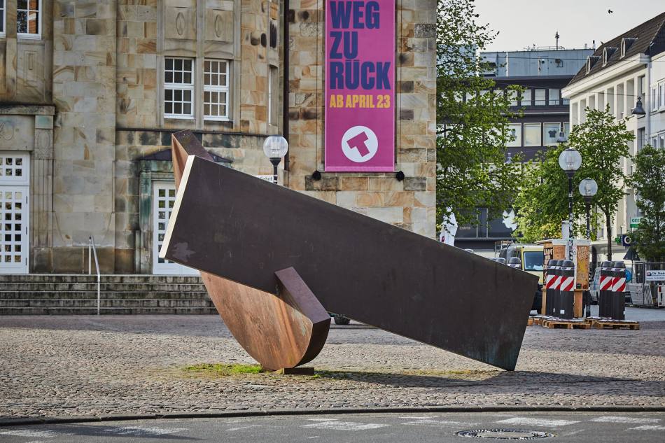 2 massive Metallelemente, die miteinander verbunden sind. Das eine hat die Form eines Halbkreises, das andere ist lang und rechteckig. Die Elemente stützen sich gegenseitig.