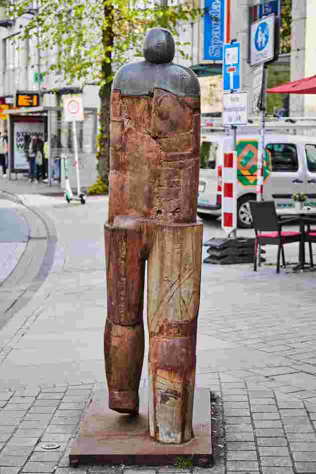 Ansicht der Skulptur von hinten: Männliche Figur, aus Eisen- und Bronzeteilen zusammengesetzt. Die Figur bewegt ihren linken Fuß nach vorne.