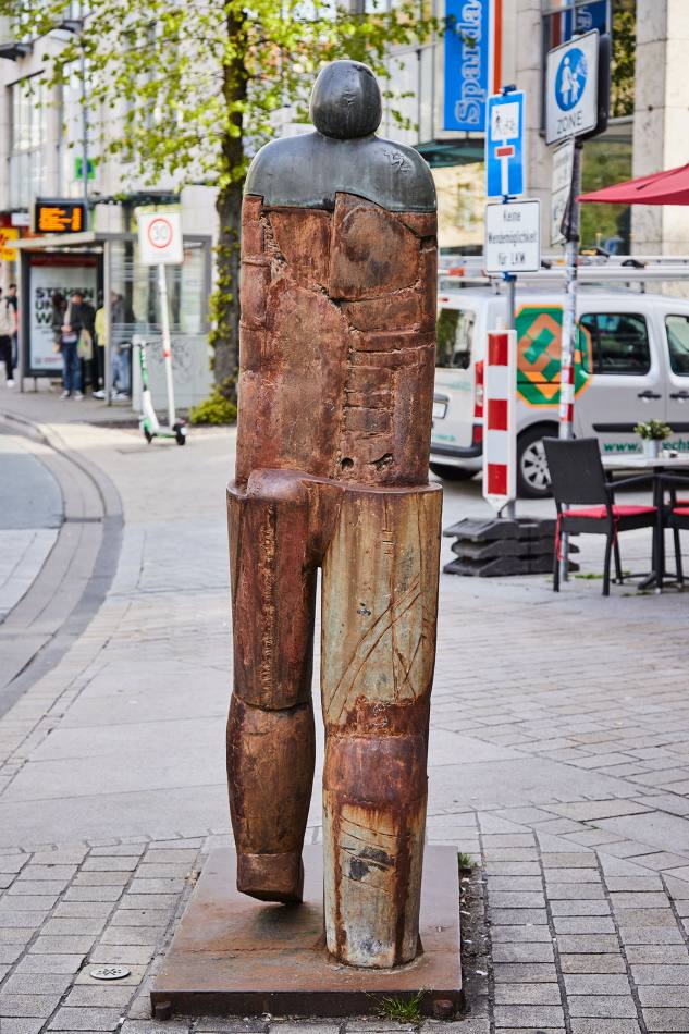 View of the sculpture from behind: Male figure, composed of iron and bronze parts. The figure is moving his left foot forwards.